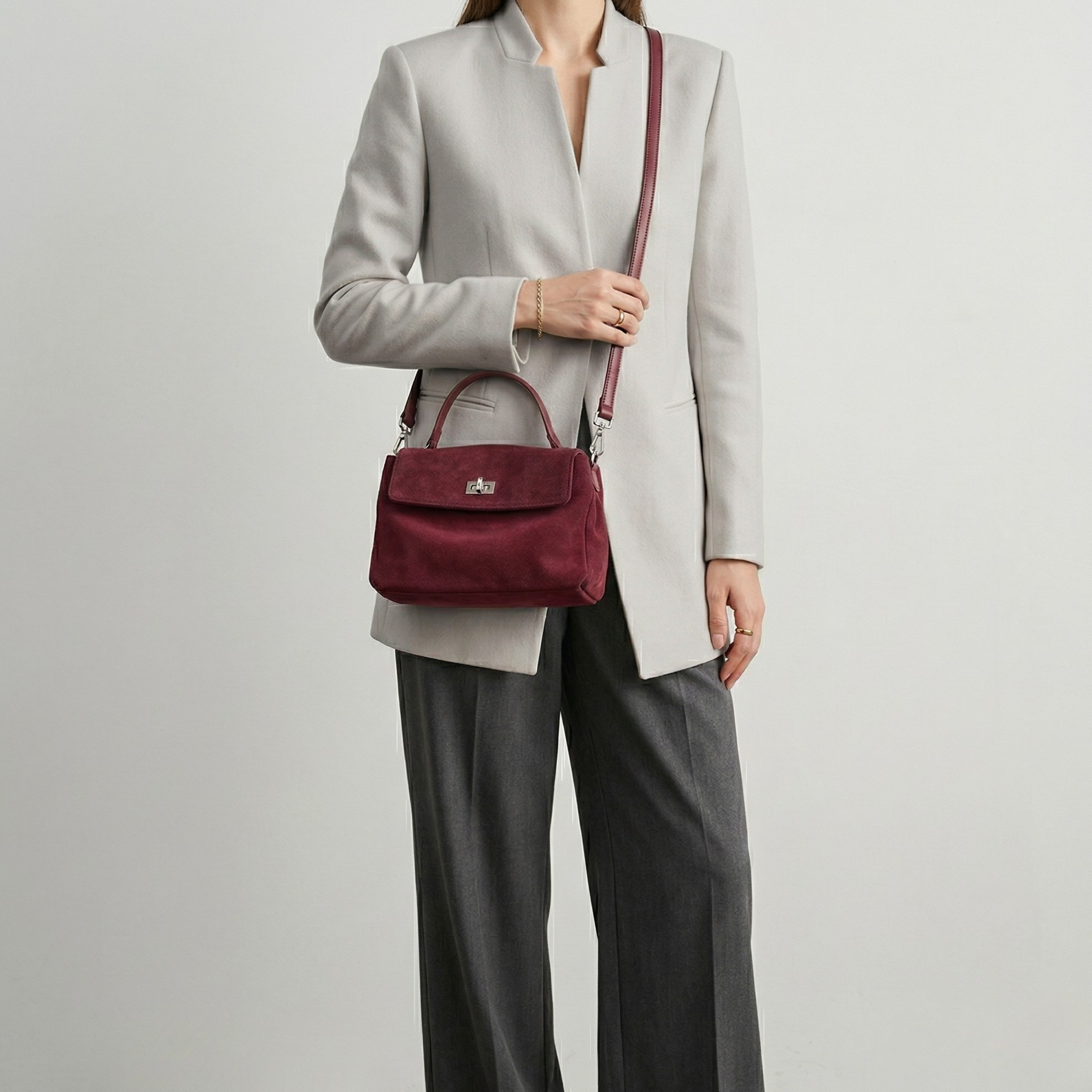Person wearing a light gray blazer and dark gray pants holding a maroon handbag against a plain background